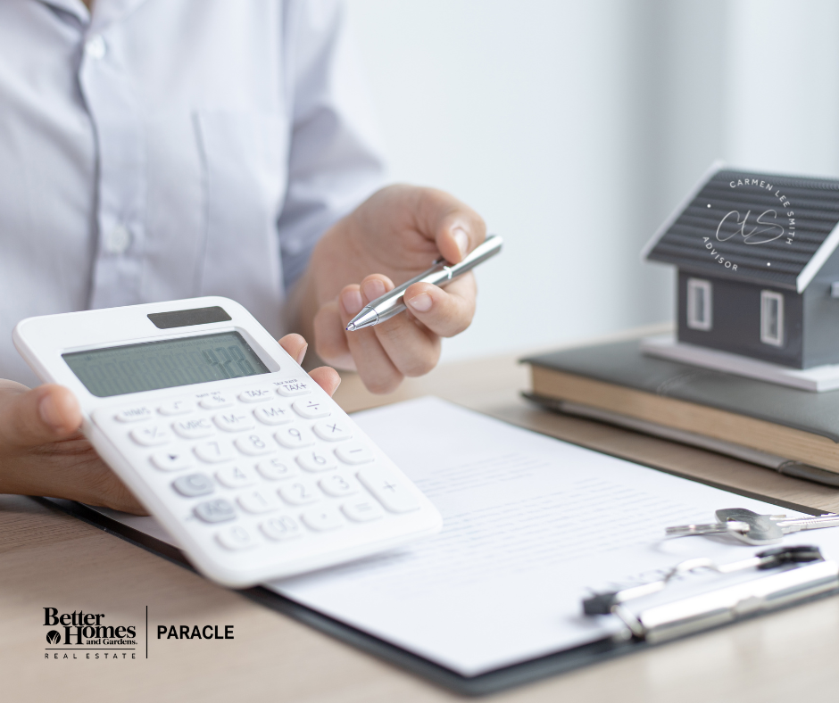 Person holding a pen in one hand and a calculator in the other is showing an off camera person who is sitting on the other side of the desk the calculator. Desk has a clip board, a notebook that has a little house on top of the book.